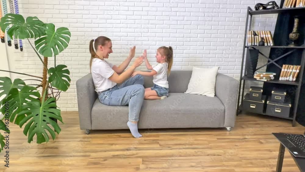 Smiling young mother and adorable little daughter clapping hands, sitting on couch, loving mum and cute toddler child girl having fun together, enjoying leisure time Smiling young mother and adorable little daughter clapping hands, sitting on couch, loving mum and cute toddler child girl having fun together, enjoying leisure time