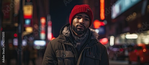 A man with a red hat and black jacket stands on a city street at night  surrounded by darkness. His facial hair is visible under the dim streetlights