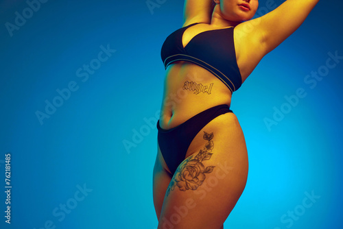 Cropped portrait of young attractive woman posing in dark bikini raising hands showing her curves in yellow neon light against gradient background. Concept of beauty, body positivity, spa procedures. © Lustre