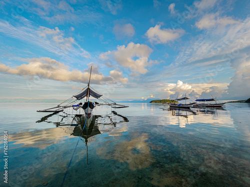 View of a fishing boat on the sea with a mirror reflection in the water against a background of blue sky with clouds shot at a wide angle (ID: 762184054)