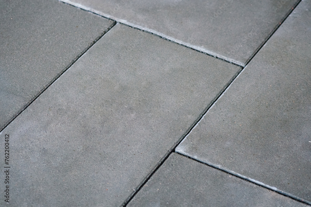 A close-up photo of a concrete sidewalk with interlocking tiles, in shades of grey and white. The texture of the concrete is visible background