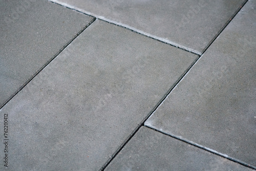 A close-up photo of a concrete sidewalk with interlocking tiles, in shades of grey and white. The texture of the concrete is visible background