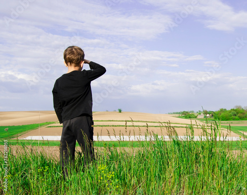 7 year old boy in black tracksuit looks in distance on background of beautiful green agricultural field.Wheat field. Agriculture.