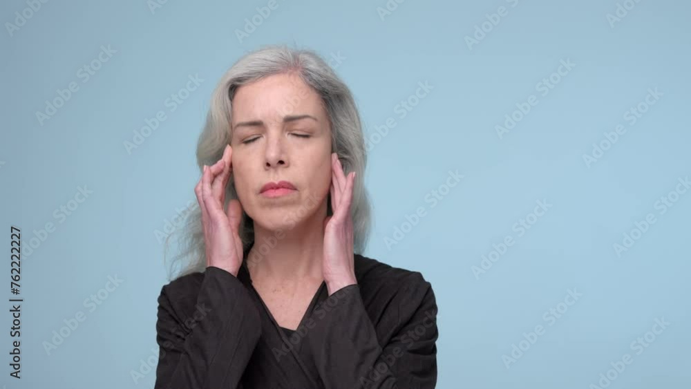 A woman with closed eyes and silver hair pressing her hands to her temples in a gesture that suggests she may have a headache, stress or want to relieve tension. A woman with closed eyes and silver hair pressing her hands to her temples in a gesture that suggests she may have a headache, stress or want to relieve tension.