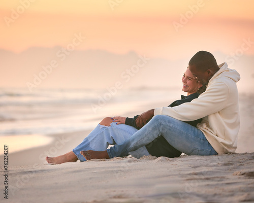 Casually Dressed Loving Young Couple Sitting On Beach Shoreline Hugging At Sunrise