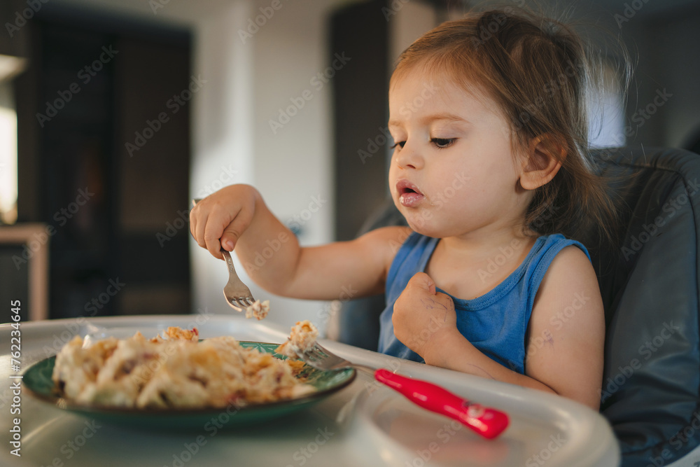 Young girl enjoying pasta in high chair with fork, Tableware