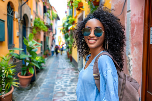 Smiling young african american woman in sunglasses travels along a colorful narrow Italian street. Summer travel concept.