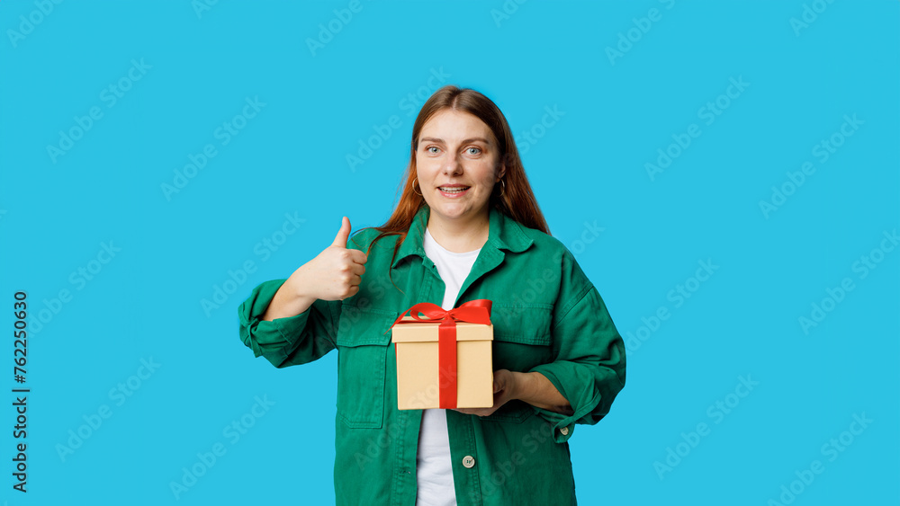Portrait of cheerful, happy 30s Girl with holding wrapped present and giving a thumb up or good luck gesture isolated on blue background, studio portrait. Portrait of cheerful, happy 30s Girl with holding wrapped present and giving a thumb up or good luck gesture isolated on blue background, studio portrait.