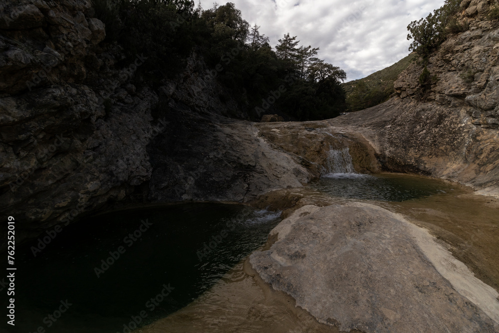 A secluded waterfall flows into a serene pond, surrounded by rocky terrain and lush greenery, evoking a sense of tranquility A secluded waterfall flows into a serene pond, surrounded by rocky terrain and lush greenery, evoking a sense of tranquility