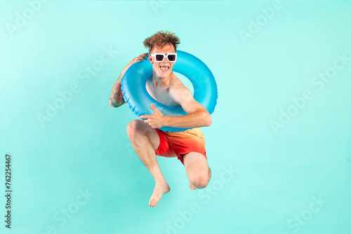 Fototapeta Naklejka Na Ścianę i Meble -  young cheerful guy winner with inflatable swimming ring jumps and flies on blue isolated background in summer