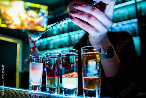 bartender skillfully pouring a layered shot at a bar. Various filled shot glasses adorn the bar, illuminated by ambient lighting. The bartender’s hands are in focus, one holding a spoon