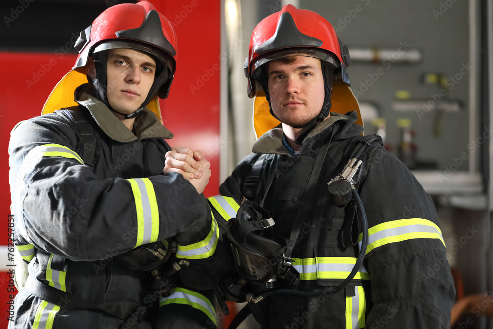 Two firefighters in protective clothing in helmets with fire engine, friendly handshake Two firefighters in protective clothing in helmets with fire engine, friendly handshake