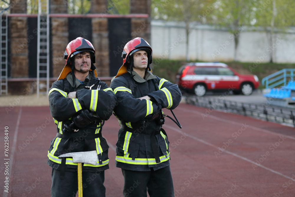 Two firefighters in equipment and red helmets on test area against fire tower stand and look at sky Two firefighters in equipment and red helmets on test area against fire tower stand and look at sky