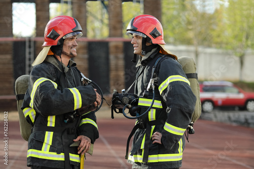 Two firefighters in equipment and helmets on test site, communicate, smile