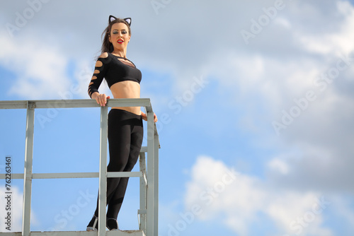 Woman dressed as black cat standing on the roof with white metal railing in front of blue sky with clouds photo