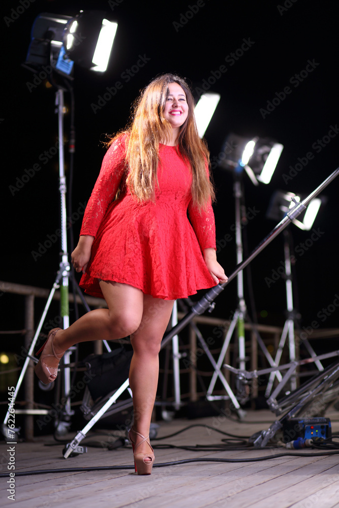 Happy plump woman with raised leg in short red dress in front of row of projectors in the dark Happy plump woman with raised leg in short red dress in front of row of projectors in the dark