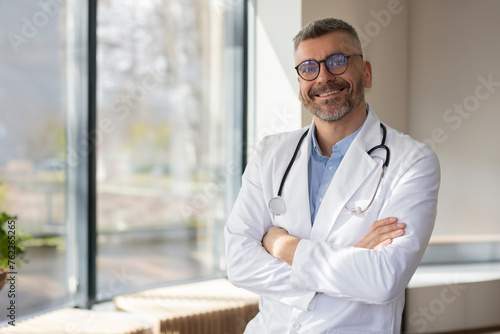 Positive middle aged european male doctor with stethoscope on neck posing with folded arms in clinic near window, looking and smiling at camera, free space