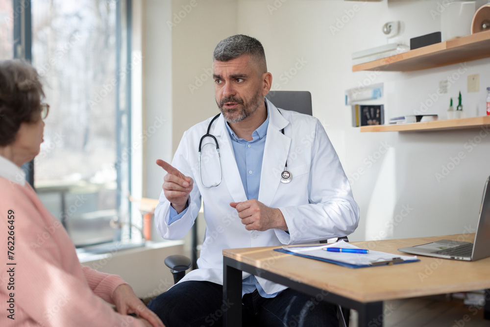 Fototapeta premium Middle aged male doctor consulting senior female patient in clinic, GP have consultation with elderly woman client in private hospital