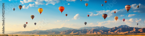 A field of colorful hot air balloons against a clear blue sky, with a banner advertising a local festival or community event. © HASHMAT