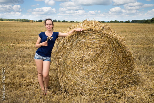 Young woman in shorts and t-shirt is standing near huge haystack.