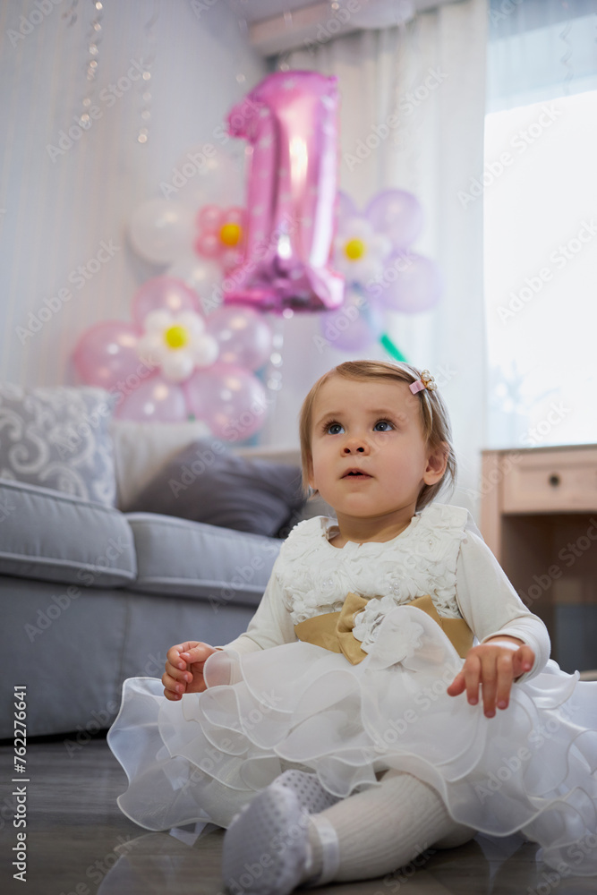 Little girl in white holiday dress sits on floor in room decorated with balloons shaped as number one for first birthday. Little girl in white holiday dress sits on floor in room decorated with balloons shaped as number one for first birthday.