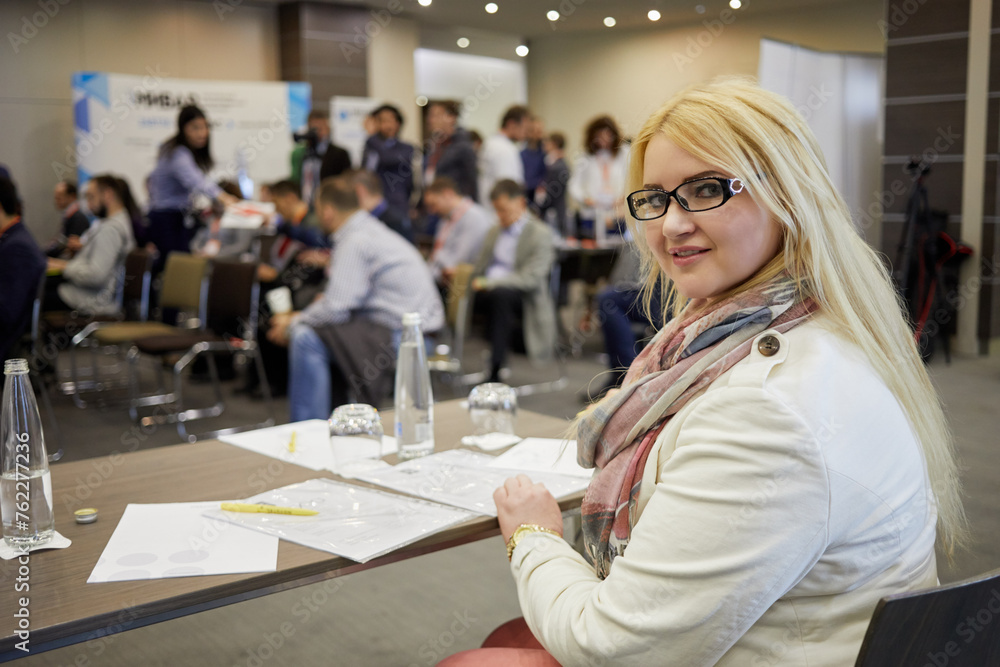 Young blond woman sits at table with papers in auditorium, shallow dof.