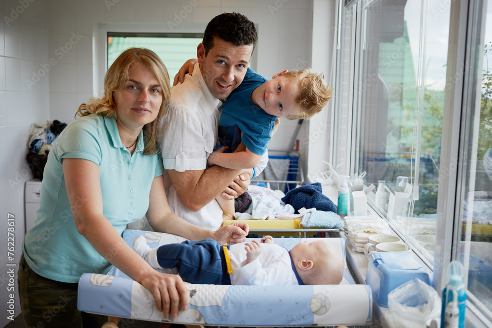 Obraz premium Smiling mother, father and son at changing table with baby lying on in room.