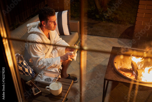Foto Man in white bathrobe sits with cigar in his hand and cup of coffee on the table in front of portable fireplace