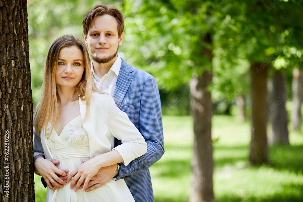 Fototapeta premium Young bearded man and woman stand ebracing at big tree trunk in park.