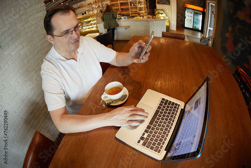 Man sits working at table with laptop, smartphone and cup of tea in cafe.