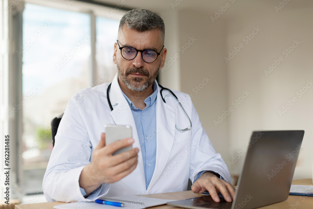 Middle aged male doctor in white uniform using cellphone and consulting patients online while sitting at desk with laptop in clinic office Middle aged male doctor in white uniform using cellphone and consulting patients online while sitting at desk with laptop in clinic office