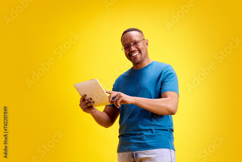 Smiling confident African-American man in glasses and blue t-shirt using tablet against yellow studio background. Freelance projects. Concept of human emotions, casual fashion, lifestyle