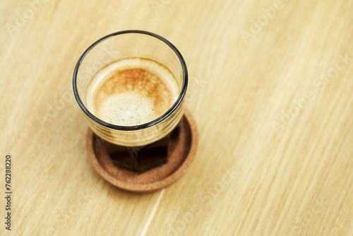Black coffee with foam in glass on saucer on wooden desk.