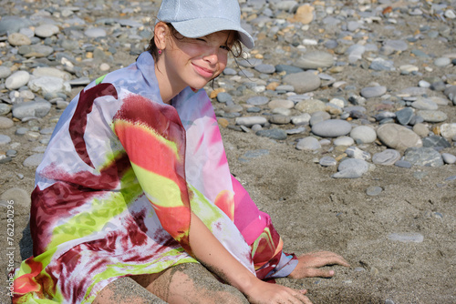 A teenage girl, wearing a cap and pareo, sits on a pebbled beach and looks into the camera.