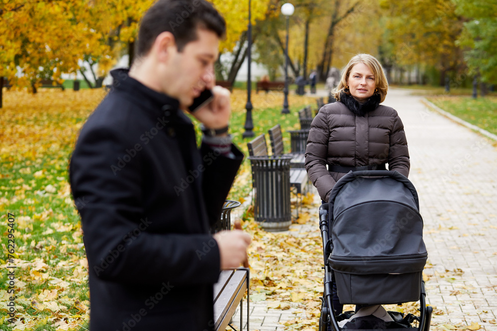 Man in profile talking cell phone and woman with perambulator in autumn park, focus on woman.