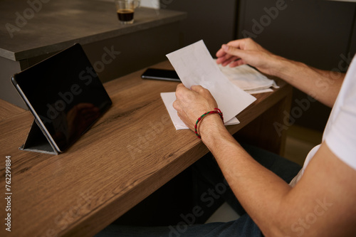 Close-up man sitting in his home office with bills in hands and paying it online over digital tablet. E-banking concept.