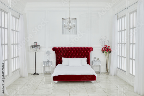 Interior of spacious white bedroom with large red bed, chandelier, lamps, flowers, windows on both sides in Ligtning Hall of Fotoelite Studio. photo