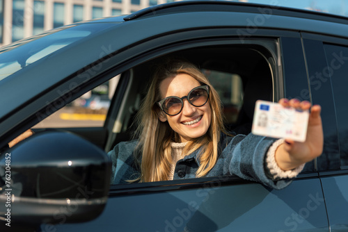 smiling young woman showing her new driver license out of car window after successful test at driving school