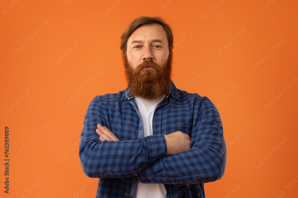 Redhaired bearded man crossing arms on chest against orange backdrop Redhaired bearded man crossing arms on chest against orange backdrop