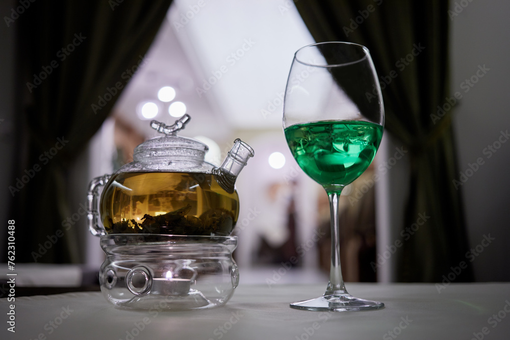Fototapeta premium Transparent glass teapot on candle heater and wineglass with green beverage with ice on table.