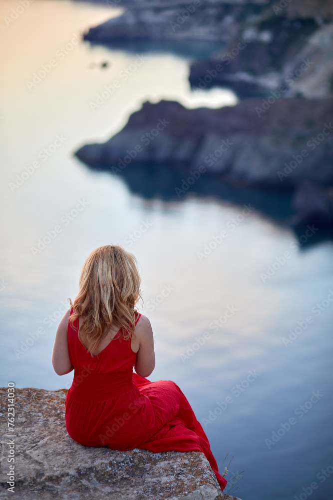 Woman in red dress sits on edge of stone cliff looking at seashore, rear view.