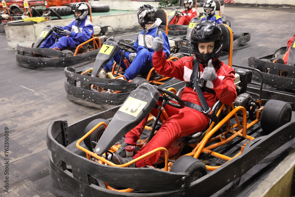 Five adults and one child in helmets prepares for driving karting, Woman in red thumbs up, focus on right woman