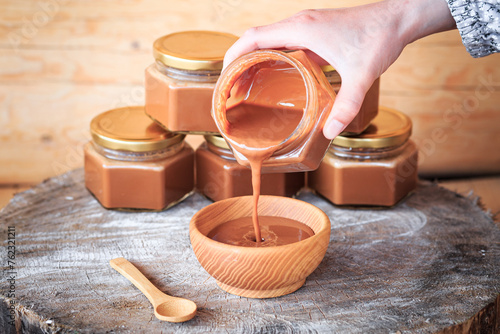 A girl's hands pour boiled condensed milk into a wooden plate, close-up against the background of glass jars filled with condensed milk, food photography, wooden background. Vertical orientation.