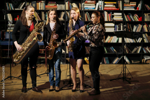 Four young pretty women with wind instruments perform in room with books