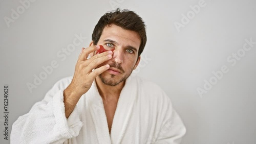 A young man in a bathrobe applies undereye patches against a white background. photo