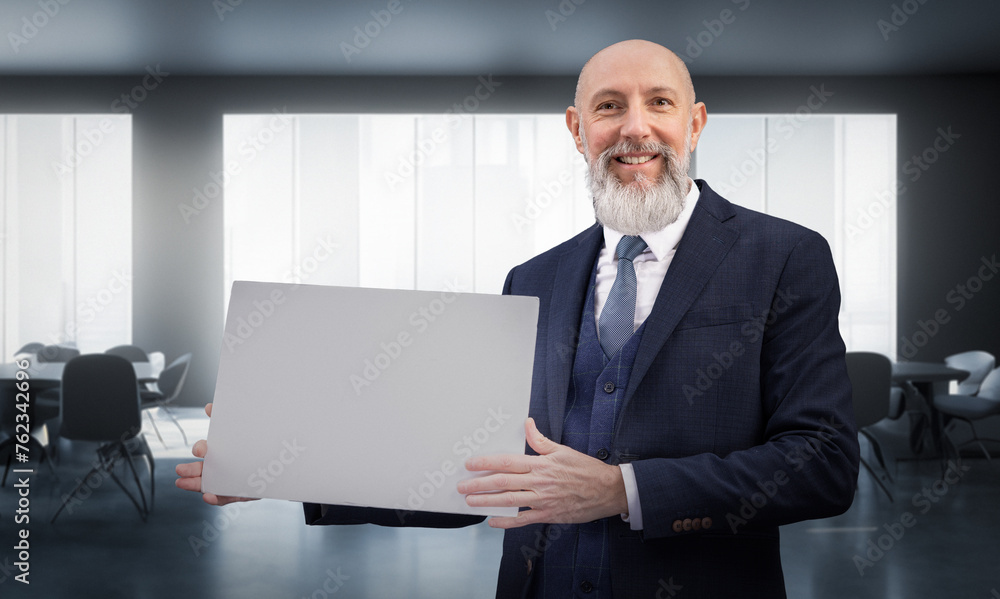 Portrait corporate d'un homme d'affaires souriant en costume et avec barbe qui tient un panneau affiche blanc dans un bureau Portrait corporate d'un homme d'affaires souriant en costume et avec barbe qui tient un panneau affiche blanc dans un bureau
