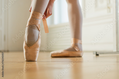 Ballerina puts on pointe shoes on leg in dance class. Classical ballet dancer woman in dance studio. Ballerina putting on tying classical pointe shoes for dance training. Ballet school concept