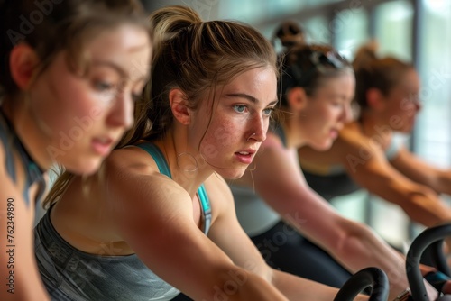 A group of women wearing workout clothes pedaling on stationary bikes in a gym setting