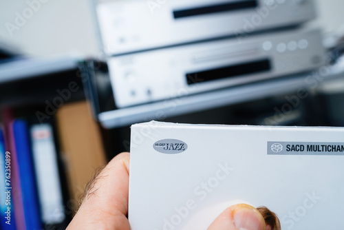 A POV shot features a male hand holding a new SACD package adorned with hi-def jazz signage, with a defocused hi-fi audiophile system in the background, setting the stage for an immersive musical photo