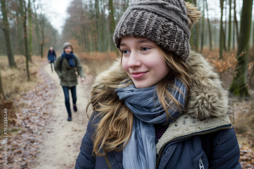 Portrait of happy young woman on forest path, wearing winter jacket and woolen hat with her mother walking in the background Portrait of happy young woman on forest path, wearing winter jacket and woolen hat with her mother walking in the background